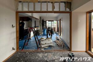 マンションのリビングとダイニングの間仕切りを解体している様子。中央で作業員が床に落ちた石膏ボードをまとめ、奥では別の作業員が脚立に上って解体作業をしている。