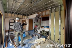 天井や壁を解体し、スケルトン状態に近づいているマンション室内。3人の作業員が脚立や手作業で壁の解体を進めている。