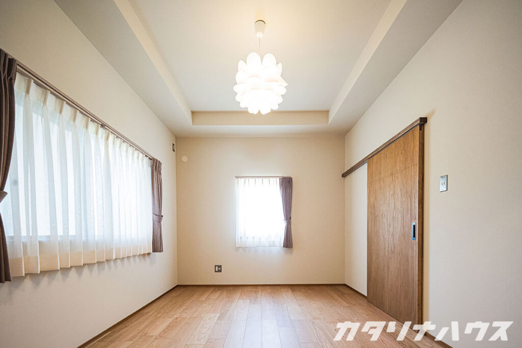 明るい洋室。二つの窓に薄いカーテンがかかり、天井から白い花のようなペンダントライトが下がるシンプルな空間。
