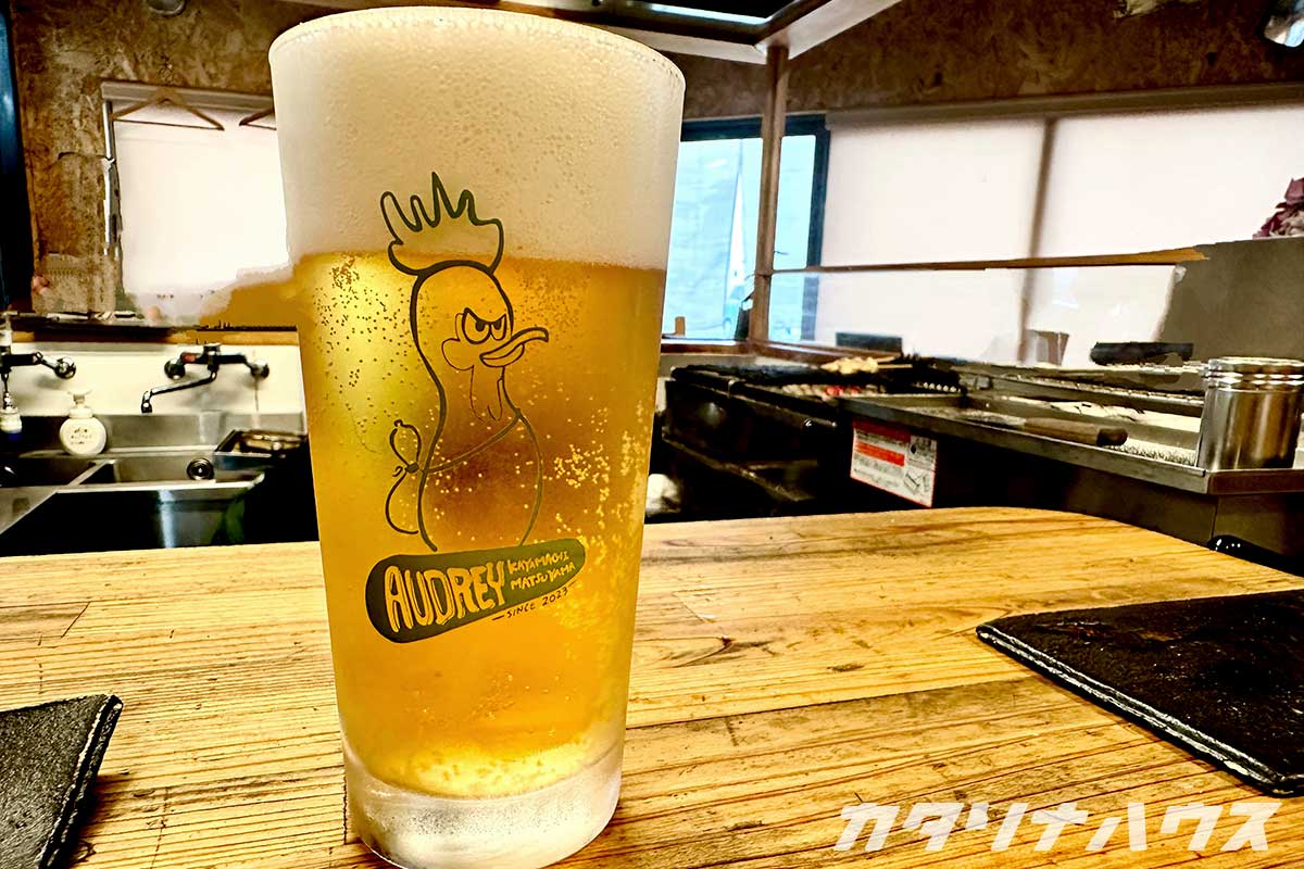 黄緑色で描かれた目つきの鋭いニワトリのイラストが描かれたグラス。泡立ったビールが入っている。