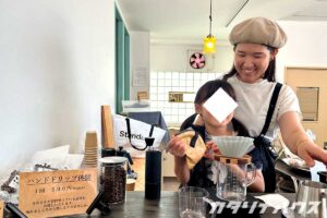 ベレー帽をかぶった女性にコーヒーの淹れ方を教わるツインテールの女の子。ハンドドリップ体験の看板が画面左に置いてある。