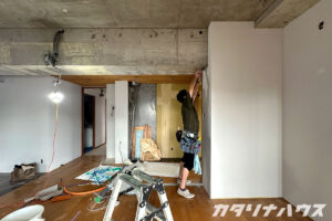職人が上に手を伸ばし、高い位置からクロスを貼る作業をしている。