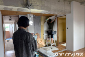壁紙を貼る職人を撮影する女性スタッフの後ろ姿。