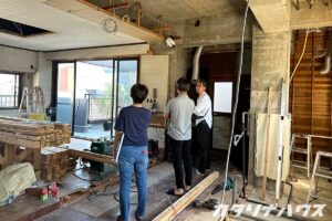 リノベーション中の室内で、解体後のコンクリートが見える空間をスタッフと施主が確認している様子。床材や木材が並べられ、設備の位置を打ち合わせしている場面。