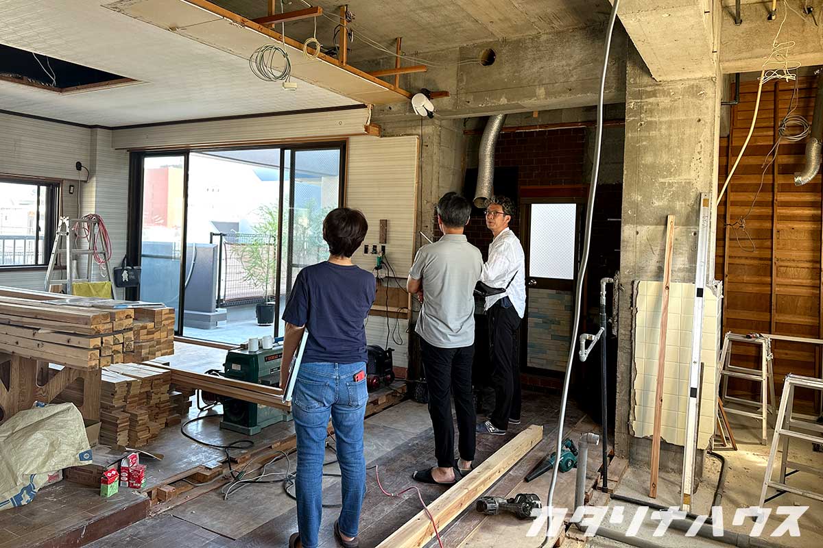リノベーション中の室内で、解体後のコンクリートが見える空間をスタッフと施主が確認している様子。床材や木材が並べられ、設備の位置を打ち合わせしている場面。