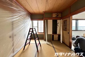 和室の壁にビニール養生が張られ、天井の配線が露出している室内。脚立が置かれ、押し入れの扉には解体予定の印が描かれているリノベーション作業中の様子。