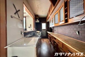 解体前の細長いキッチンで、壁に撤去予定の印が付けられ、吊り戸棚やカウンターが残るリノベーション前のキッチン空間。配線や設備の外し作業が進む様子。