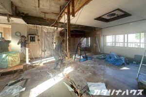 天井や壁の解体が進み、配線が多数露出した室内で職人が作業するリノベーション現場。床材の撤去や養生シートが敷かれ、スケルトン化が進む施工途中の様子。