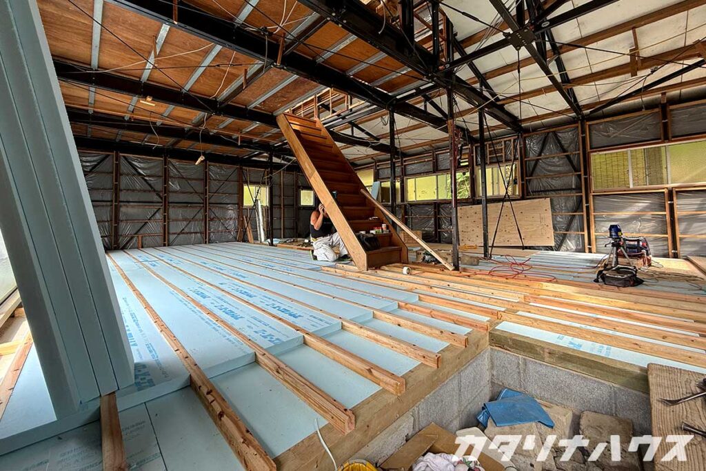 中古住宅のスケルトンリノベーション現場。床下に断熱材を敷き込む作業中で、階段の組み立てと構造体の補強が進む室内