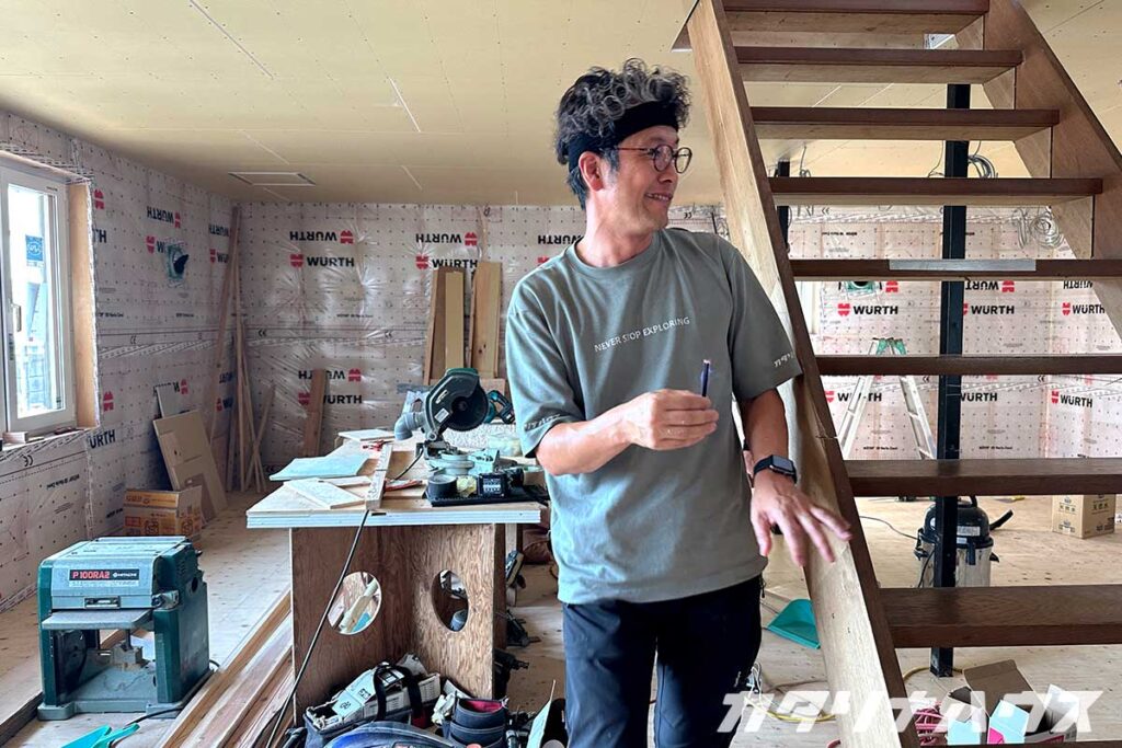 中古住宅リノベーションの室内で作業する大工さん。壁の断熱シート施工が進み、階段や作業台が設置された現場の様子