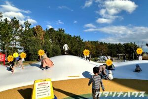 白いふわふわドーム状の大型遊具で、子どもたちが跳んだり滑ったりして遊べる公園のアクティビティエリア。青空の下、多くの家族連れでにぎわう広場。