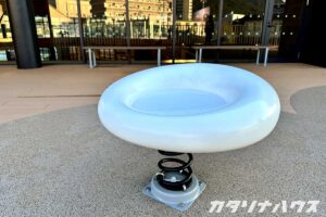 白い円形のスプリング遊具が設置されたテラススペースの一角。建物前の広場にある子ども向け遊具で、シンプルなデザインが特徴の休憩・遊びエリア。