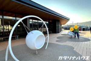 テラスに並ぶ白い椅子型ブランコとスプリング遊具が映る、開放的な遊び場スペース。木目調デッキとガラス張りの建物が並ぶモダンな外観の施設前エリア。