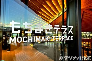 ガラス扉に「もちまきテラス」のロゴが大きく映り、館内の木の天井や明るい照明が反射する施設入口の様子。モダンな建築デザインが特徴の観光スポット。