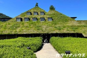 丘のように見える草屋根の斜面に、小さな窓が規則的に並ぶ建物。木々の緑と屋根の芝が連続し、自然の地形と溶け込むデザイン